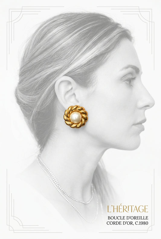 A close-up of a person's ear wearing a gold flower-shaped stud earring with a white faux-pearl center, with blonde and gray hair framing the ear.
