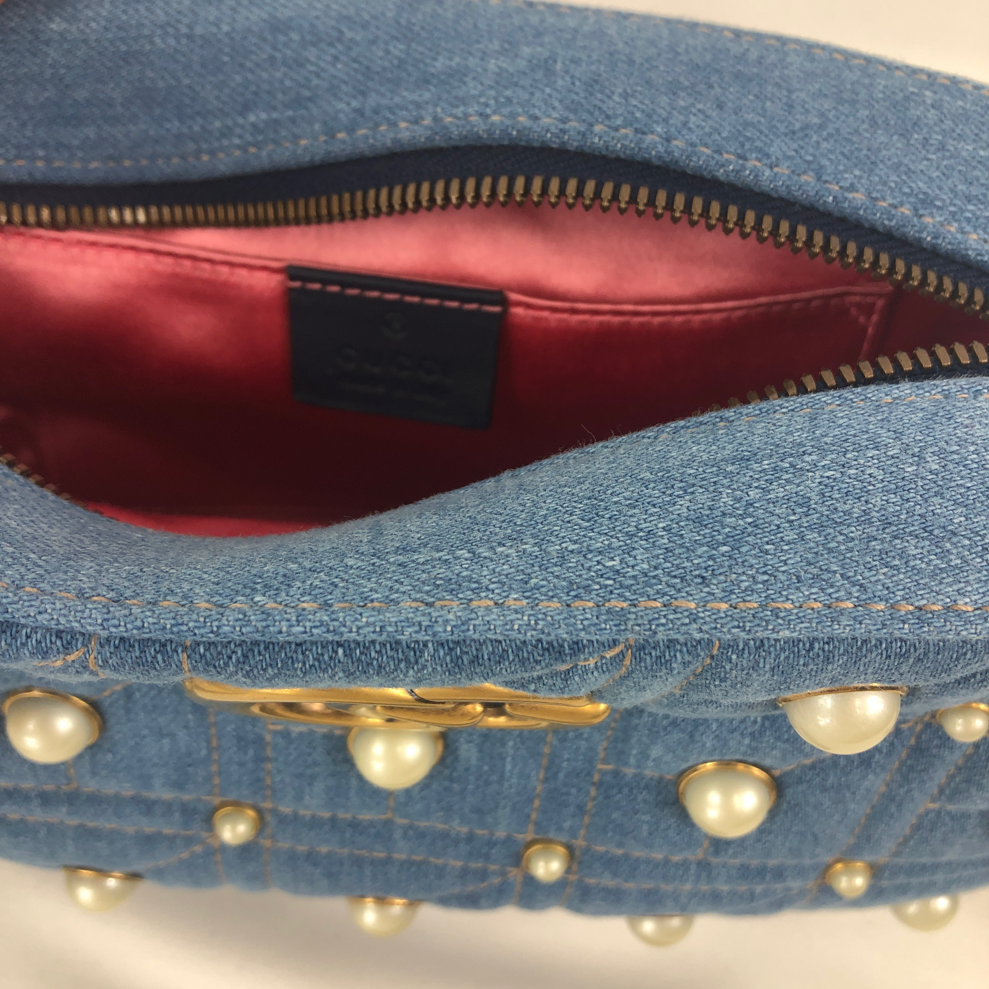 The Gucci Marmont Pearl Crossbody Bag in Denim is a modern take on a classic look. Light wash Matelassé denim Gucci Small GG Marmont Pearl Shoulder Bag with gold-tone hardware, single flat shoulder strap with chain-link accents, creme contrast stitching throughout, Running GG adornment at front face, pearl embellishments throughout exterior, coral satin interior lining, single interior slit pocket and zip closure at top. Includes dust bag. Authentic and rare handbags from Gavriel.us