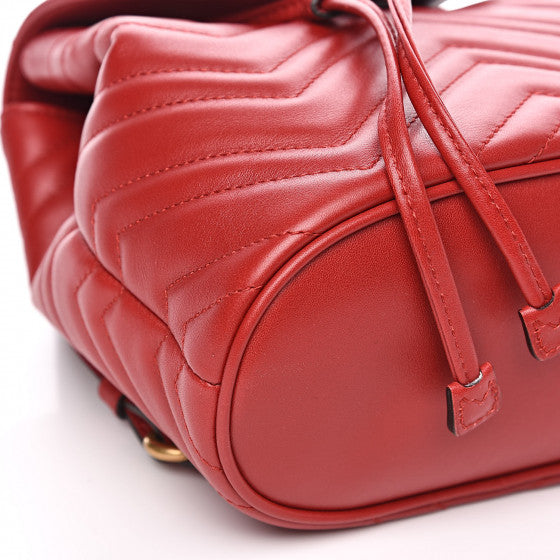 The Gucci Black Marmont Quilted Leather Backpack in Red features adjustable shoulder straps with post-stud fastening. Logo plaque at face. Fold over flap with magnetic tab fastening. Drawstring fastening at throat. Patch pocket and leather logo flag at interior. Leather lining in beige. Antiqued gold-tone hardware. Tonal stitching.