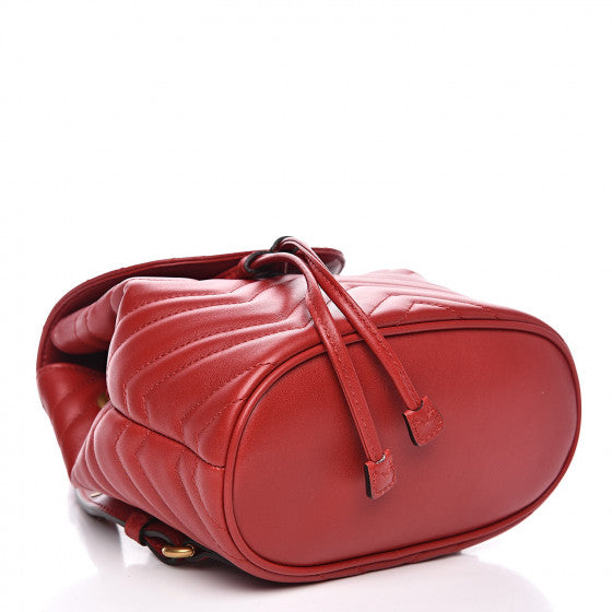 The Gucci Black Marmont Quilted Leather Backpack in Red features adjustable shoulder straps with post-stud fastening. Logo plaque at face. Fold over flap with magnetic tab fastening. Drawstring fastening at throat. Patch pocket and leather logo flag at interior. Leather lining in beige. Antiqued gold-tone hardware. Tonal stitching.