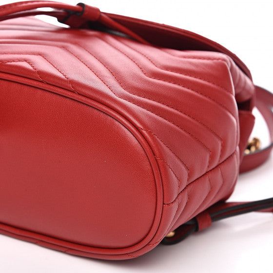 The Gucci Black Marmont Quilted Leather Backpack in Red features adjustable shoulder straps with post-stud fastening. Logo plaque at face. Fold over flap with magnetic tab fastening. Drawstring fastening at throat. Patch pocket and leather logo flag at interior. Leather lining in beige. Antiqued gold-tone hardware. Tonal stitching.