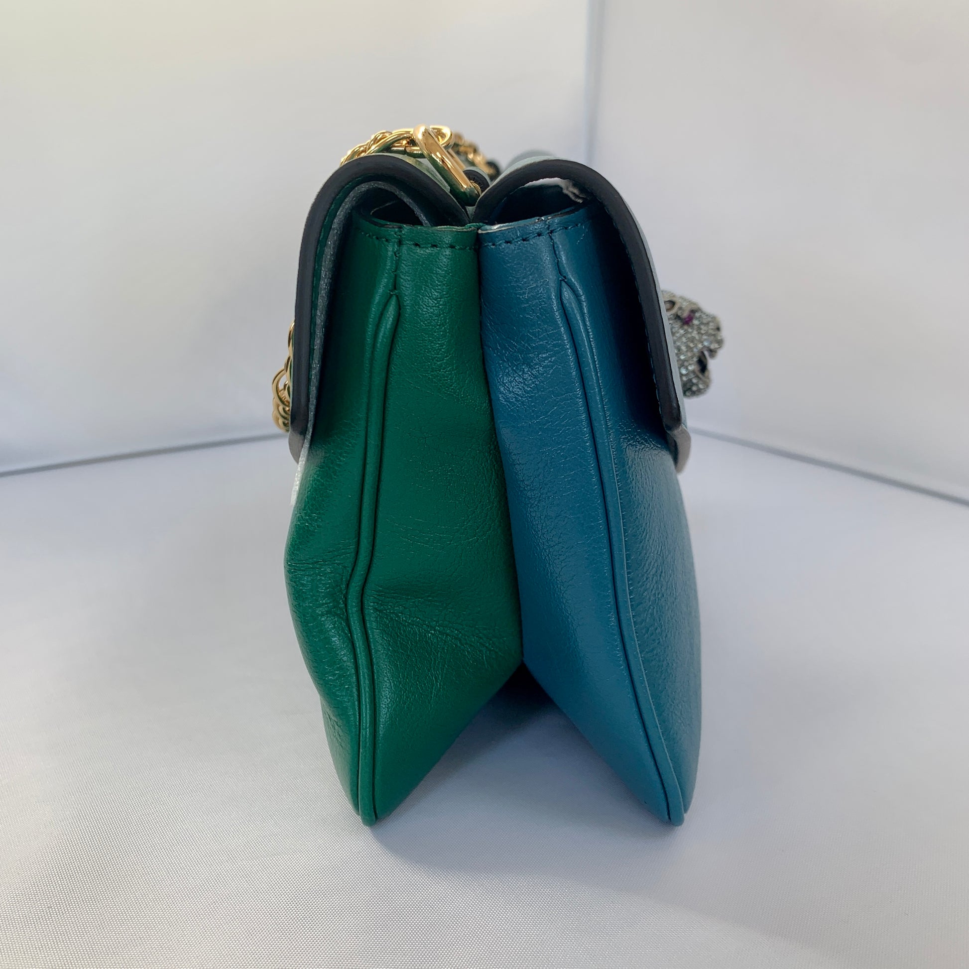 This is a two toned, double sided Gucci bag. The style is called Thiara and it looks like two envelopes back to back. One side is a mellow Robin blue. The other side is a jade green. The blue side has a jewel encrusted Raja tiger head on the magnetic clasp. The green side has the iconic double GG on the button clasp. There is a fixed Gucci gold short chain on the top and a strap is also included to make this a should bag or crossbody.