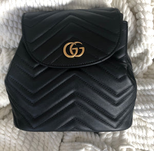 The Gucci Black Marmont Quilted Leather Backpack features adjustable shoulder straps with post-stud fastening. Logo plaque at face. Fold over flap with magnetic tab fastening. Drawstring fastening at throat. Patch pocket and leather logo flag at interior. Leather lining in beige. Antiqued gold-tone hardware. Tonal stitching.