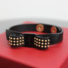 Load image into Gallery viewer, Bracelets can spice up any outfit, and this classic black, studded accessory will do just that. Soft leather was elegantly shaped to create a vara bow, which features stylish gold studs and, of course, the classic Ferragamo name. Adjustable to fit your wrist perfectly, this bracelet is meant to be shown off!