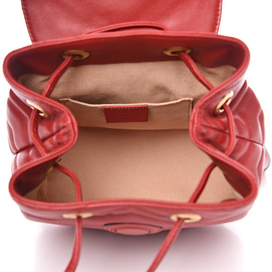 The Gucci Black Marmont Quilted Leather Backpack in Red features adjustable shoulder straps with post-stud fastening. Logo plaque at face. Fold over flap with magnetic tab fastening. Drawstring fastening at throat. Patch pocket and leather logo flag at interior. Leather lining in beige. Antiqued gold-tone hardware. Tonal stitching.