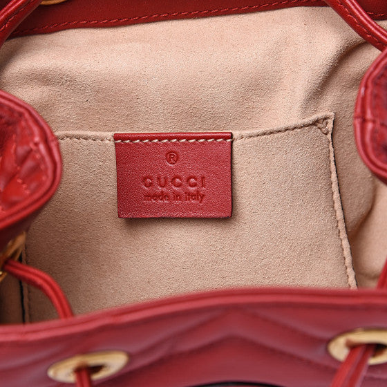 The Gucci Black Marmont Quilted Leather Backpack in Red features adjustable shoulder straps with post-stud fastening. Logo plaque at face. Fold over flap with magnetic tab fastening. Drawstring fastening at throat. Patch pocket and leather logo flag at interior. Leather lining in beige. Antiqued gold-tone hardware. Tonal stitching.