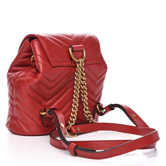 The Gucci Black Marmont Quilted Leather Backpack in Red features adjustable shoulder straps with post-stud fastening. Logo plaque at face. Fold over flap with magnetic tab fastening. Drawstring fastening at throat. Patch pocket and leather logo flag at interior. Leather lining in beige. Antiqued gold-tone hardware. Tonal stitching.